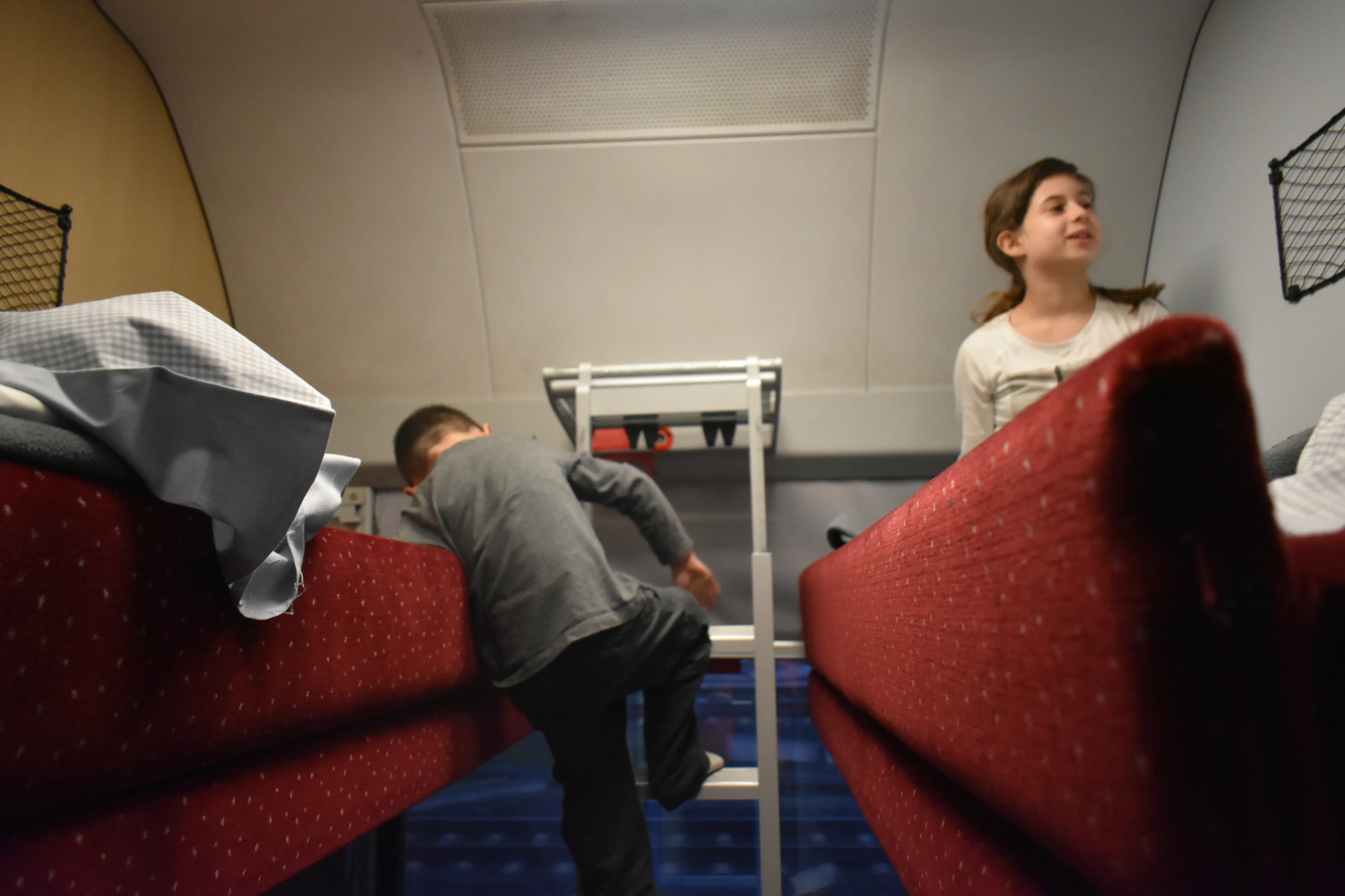 A young boy climbing a ladder to an upper bunk in a sleeper compartment on an ÖBB Nightjet train in Hamburg, while his sister sits on the other bunk.