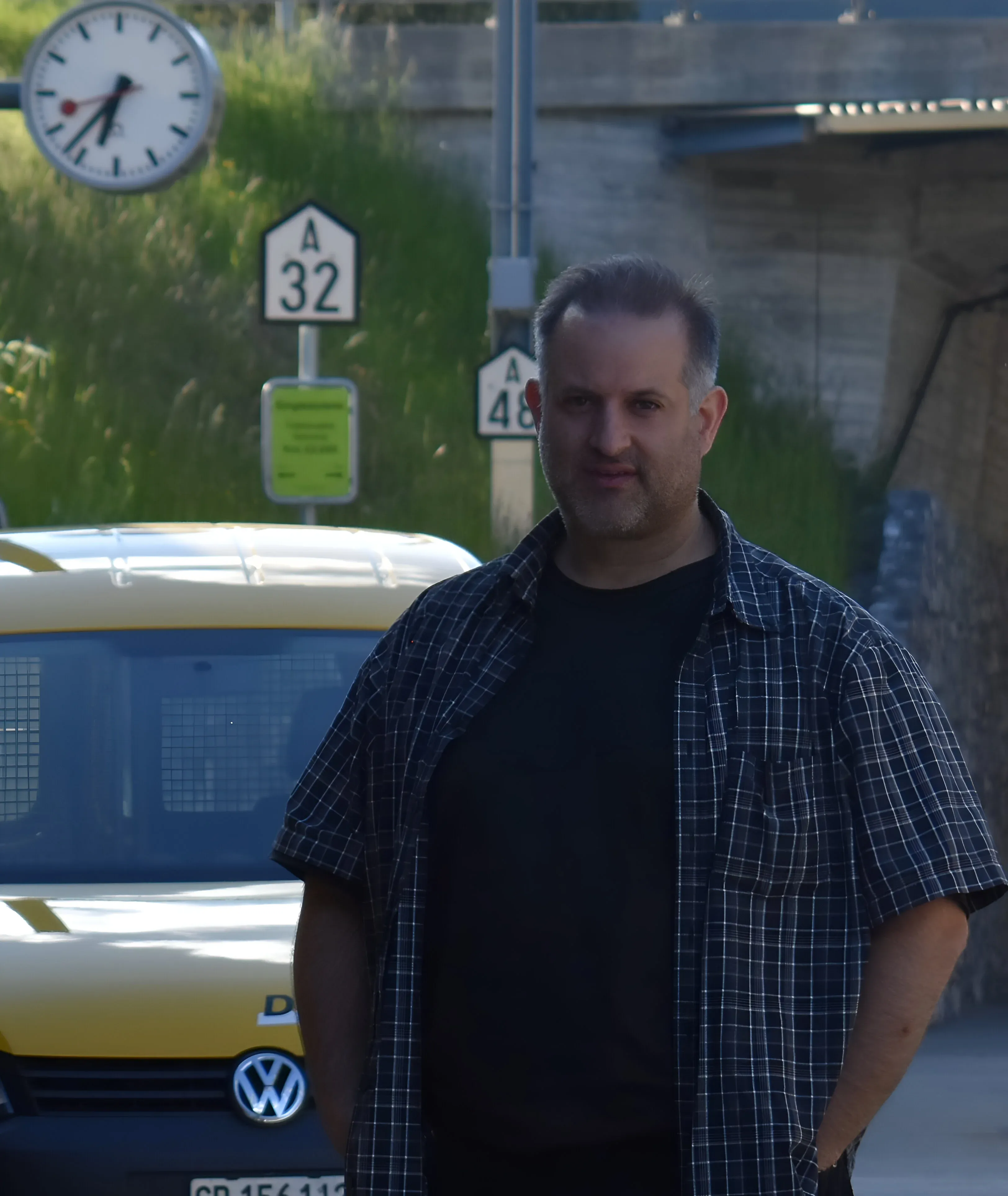 Mike from Rail Motus standing on a train platform in Tiefencastel, Switzerland. A yellow Post vehicle and station clock are in the background.