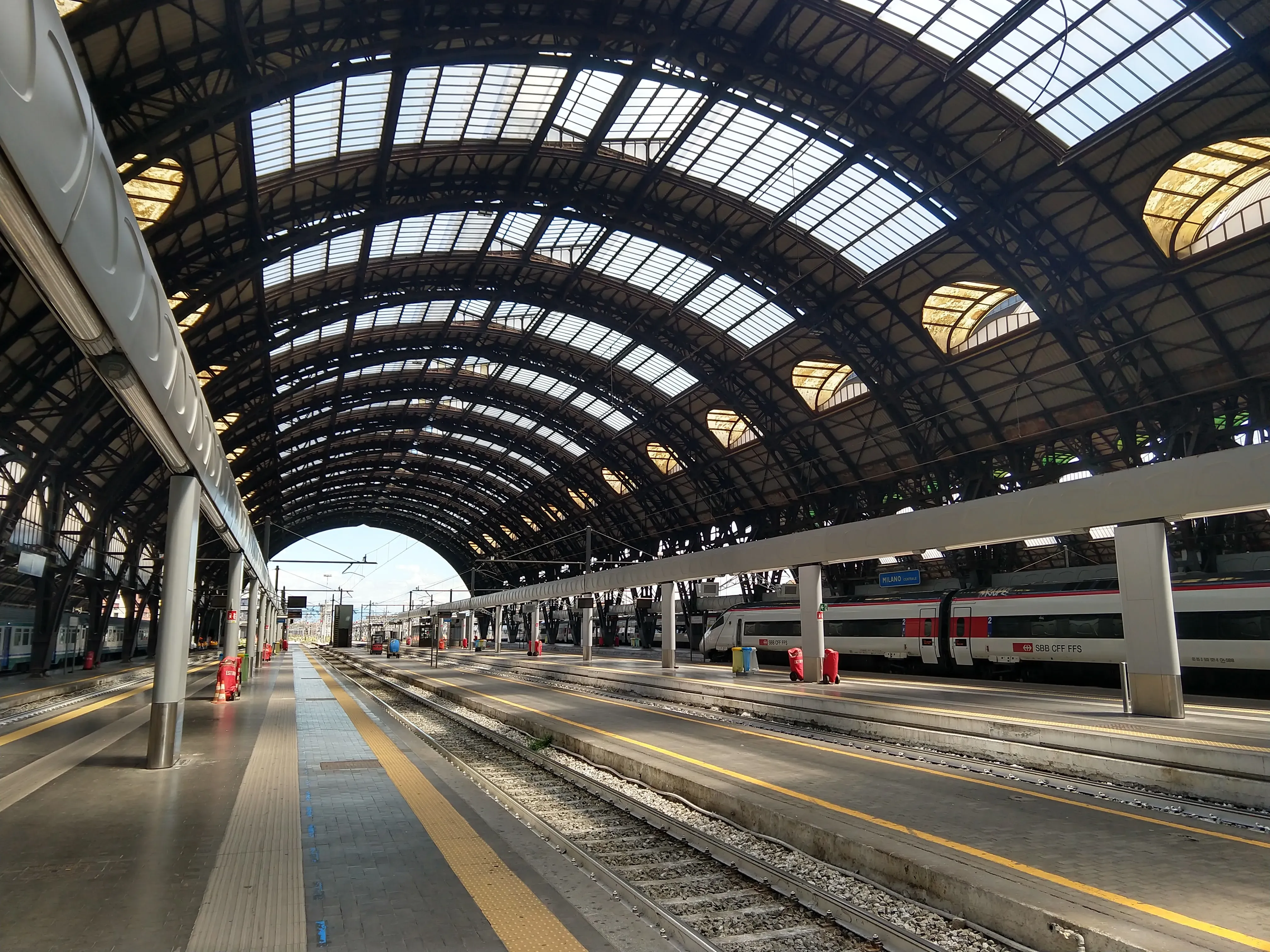 Expansive arched glass roof of Milano Centrale station, with an SBB train parked at one of the many platforms below.