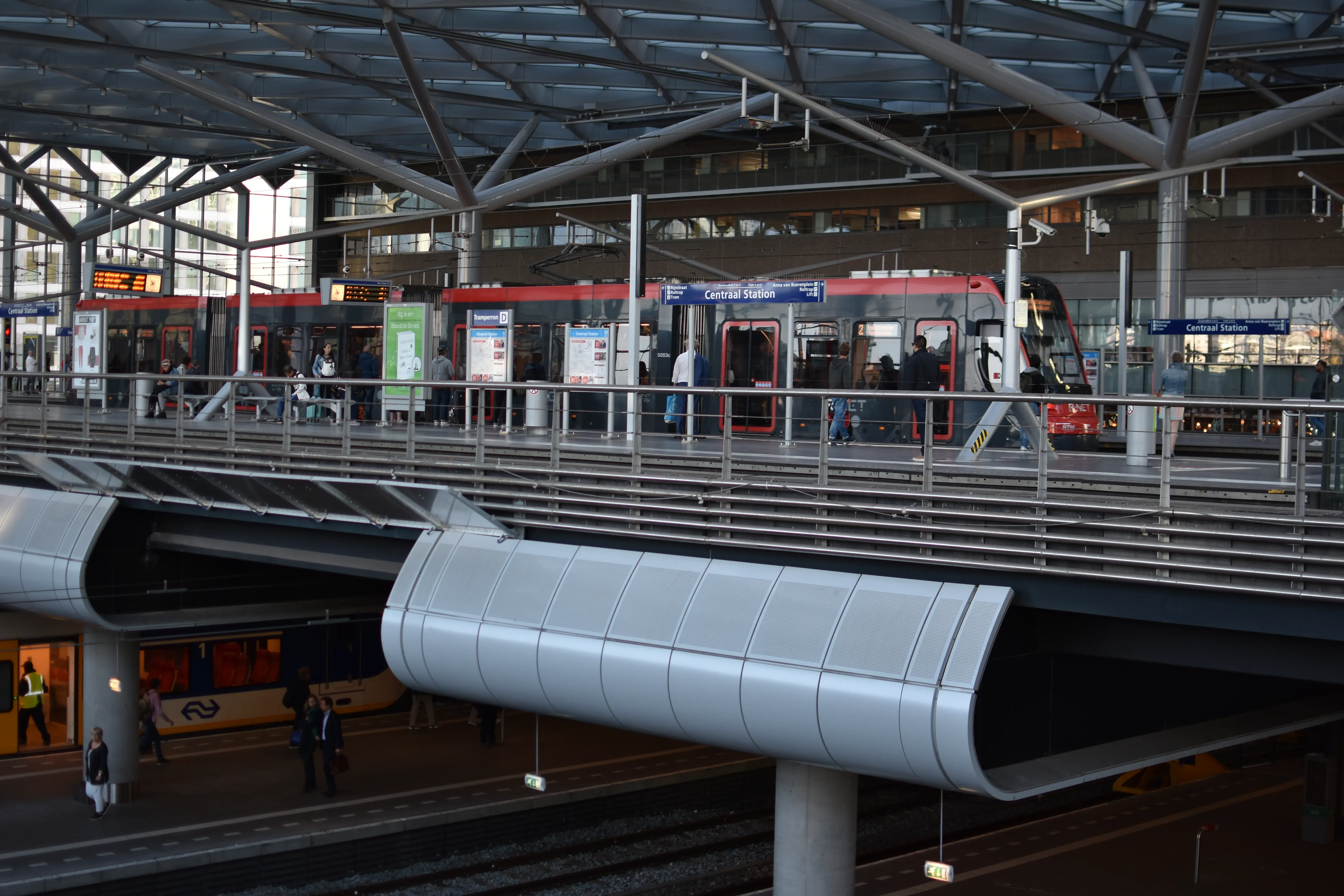 Elevated tram platform crossing over train tracks inside the glass-roofed The Hague Central Station, with a red tram waiting for passengers.